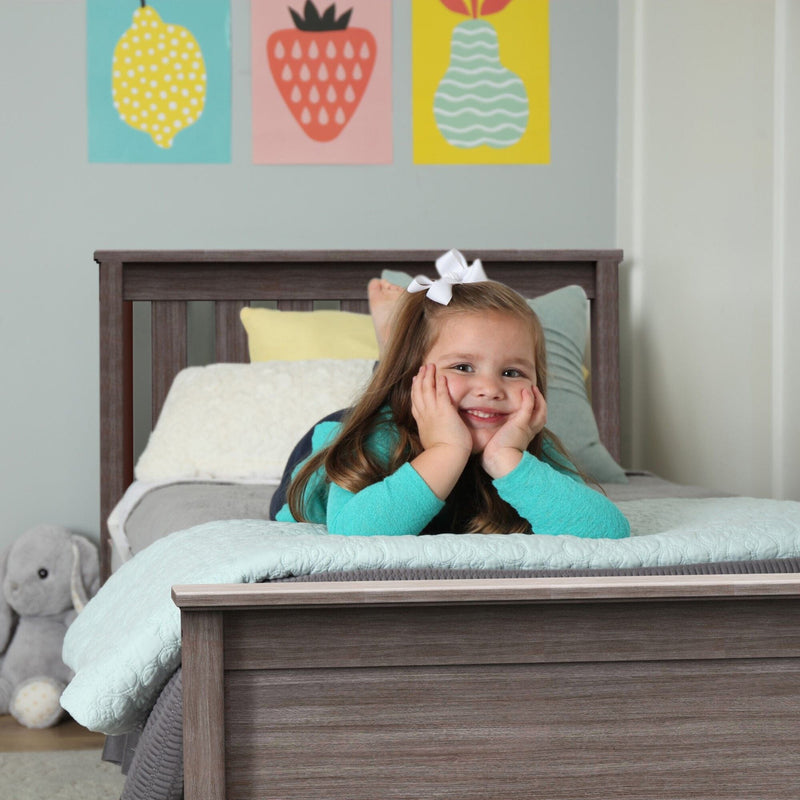 Children's bedroom decor: smiling girl lying on aqua quilted bedding on a slatted wooden bed frame with pastel fruit wall art and plush elephant toy