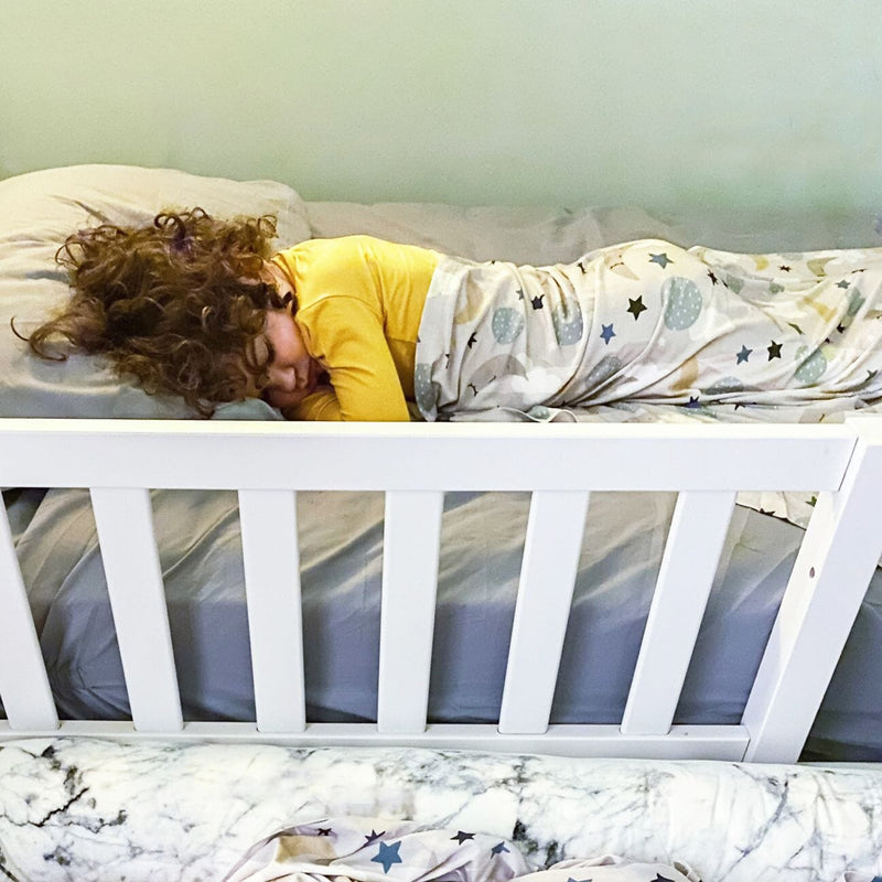 Sleeping child in yellow pajamas wrapped in star-and-moon blanket on a white-rail toddler bed — cozy nursery kids bedding