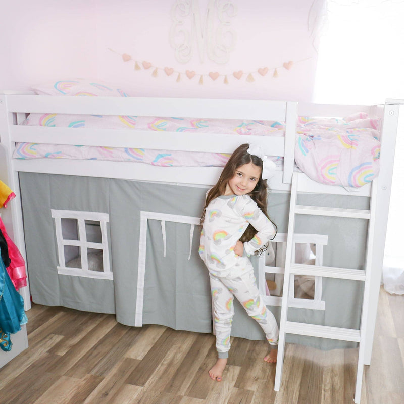 Kids loft bed with white ladder and playhouse curtain, little girl in rainbow pajamas posing beside pastel rainbow bedding and pink bedroom decor