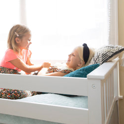 Two smiling young girls playing on a white daybed in a bright sunlit bedroom, wearing floral dresses with teal pillows and cozy knit blanket