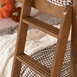 Close-up of a solid wooden bunk bed ladder with brown slatted steps in a cozy kids' bedroom featuring a checkered blanket and plush pumpkin decor