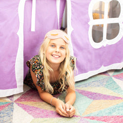 Young girl smiling while playing under a purple play tent, wearing a floral dress and a sleep mask.