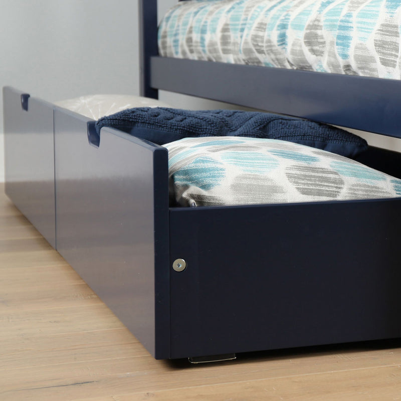 Navy blue wooden trundle bed with pull-out underbed storage drawers holding blue patterned pillows and blankets on light hardwood floor