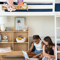 Girls reading a book under a loft bed in a stylishly decorated bedroom with a bookshelf and decor items.
