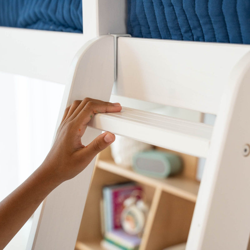 Hand holding a white wooden ladder rung of a bunk bed with blue bedding and a bookshelf in the background.