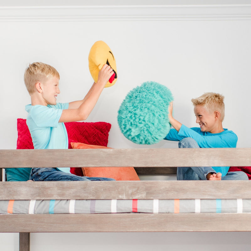 Two boys pillow fighting on a wooden bunk bed with colorful throw pillows and an emoji cushion in a bright kids' bedroom