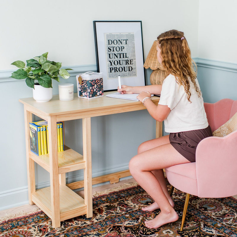 Girl studying at a light wood desk in a cozy home study nook with pink chair, potted plant, motivational "Don't Stop Until You're Proud" poster and patterned rug.