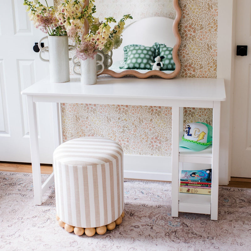 White vanity desk with scalloped mirror and striped round ottoman against floral wallpaper, styled with vases, books and a green cap — kids bedroom decor