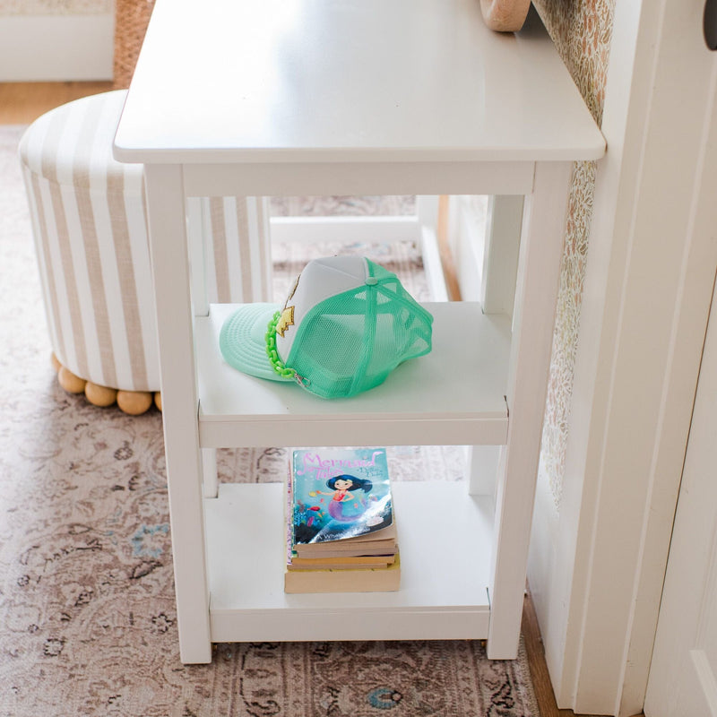 White bedside table with a mint green mesh trucker hat on the middle shelf and stacked children's books on the lower shelf beside a striped ottoman and patterned rug, cozy nursery decor