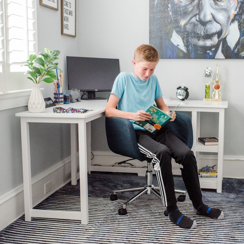 Teen boy reading a science book in a bright modern home study with a white desk, computer monitor, potted plant, trophies and striped rug