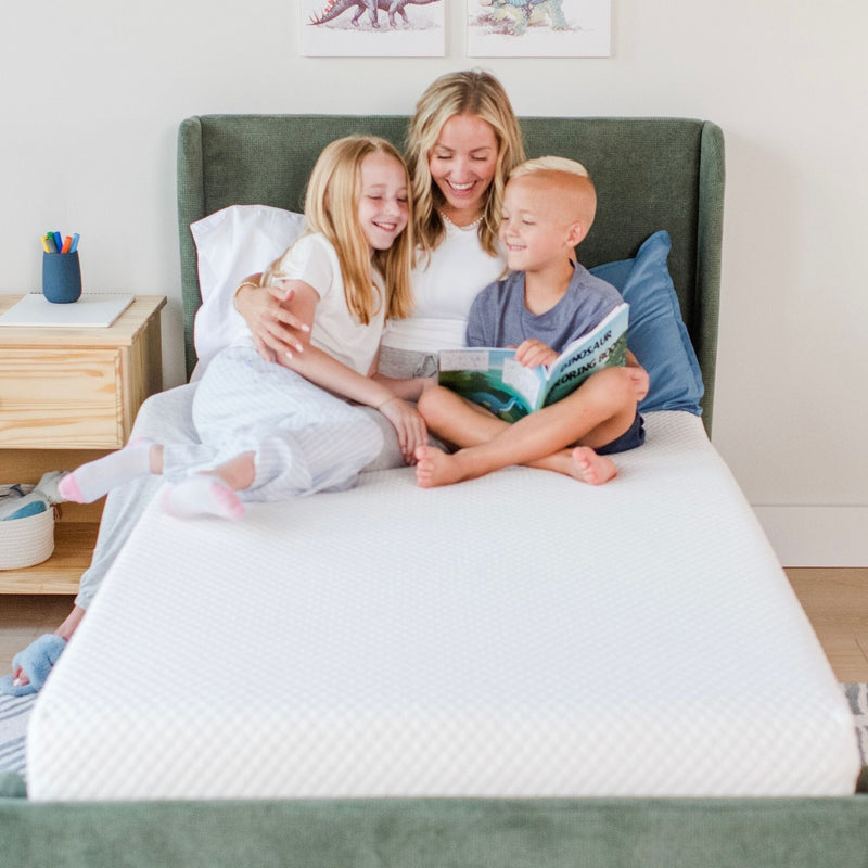Mom reading a dinosaur bedtime story to her smiling daughter and son on a white mattress in a cozy modern kids' bedroom, family bonding and bedtime routine