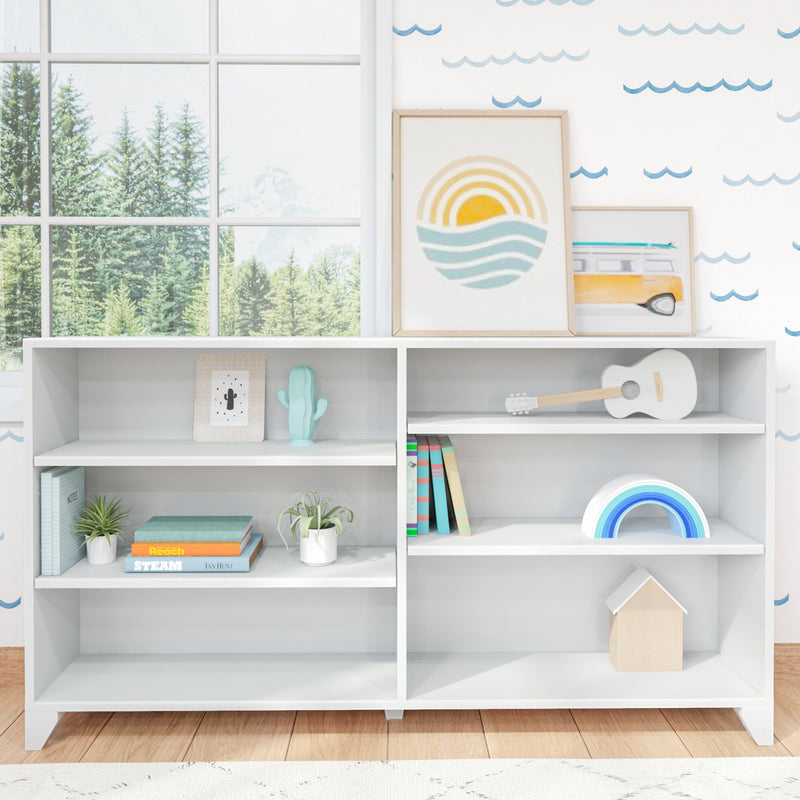 White modern kids bookshelf in nursery with pastel wooden toys, rainbow stacker, small guitar, children's books, potted plants and framed coastal art by a large window with forest view