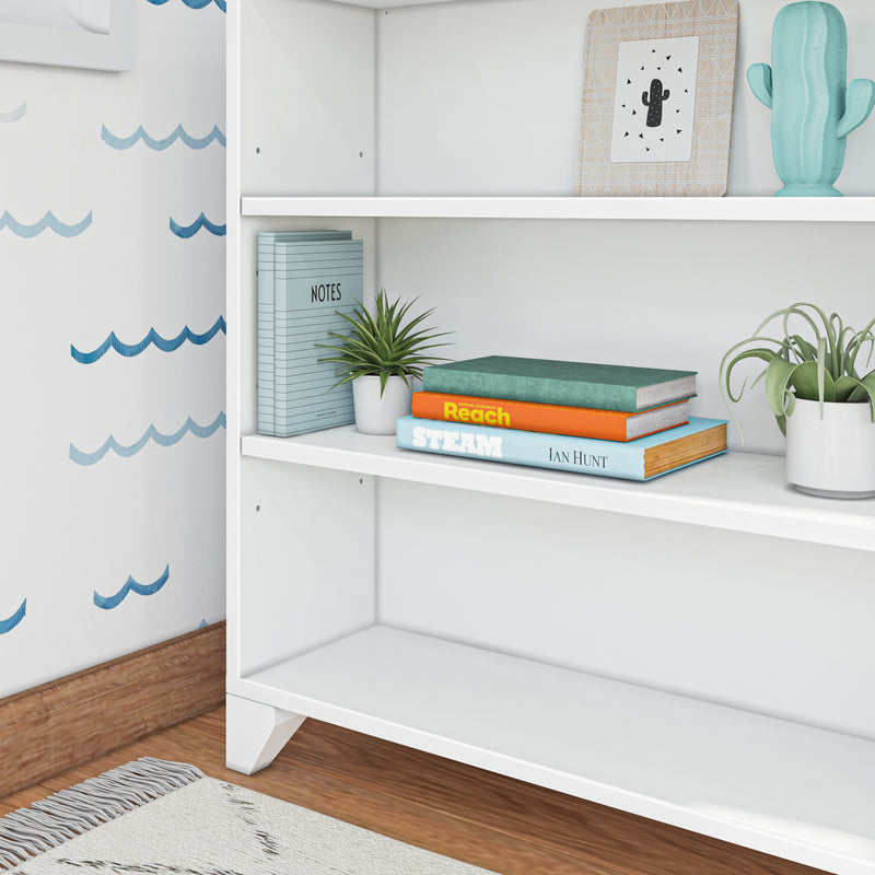 White bookshelf in a children's room with stacked educational books, potted succulents, cactus decor and blue wave wallpaper