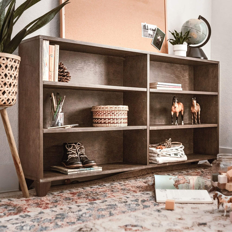 Wooden cubby bookshelf styled with woven baskets, leather boots, toy horse figurines, a globe and potted plant on a patterned boho rug