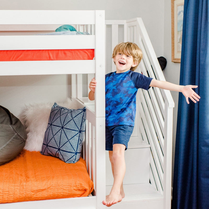 Smiling young boy playing on a white bunk bed with stairs in a modern boys bedroom with orange quilt, patterned pillows and navy curtains