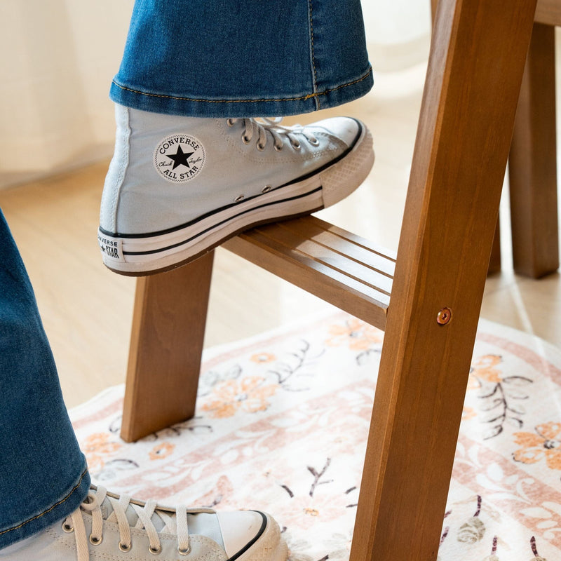 Close-up of white Converse Chuck Taylor high-top sneaker with blue jeans resting on a wooden step stool over a floral-patterned rug