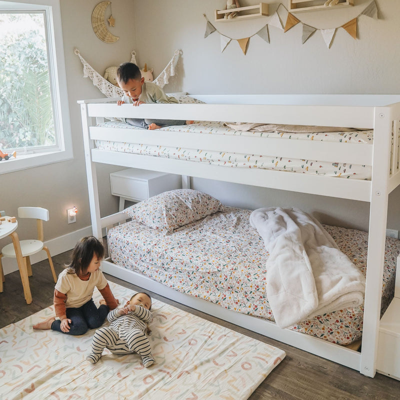 Children's bedroom with white bunk bed and floral bedding, neutral boho nursery decor and bunting, natural light from window and two kids playing on floor mat