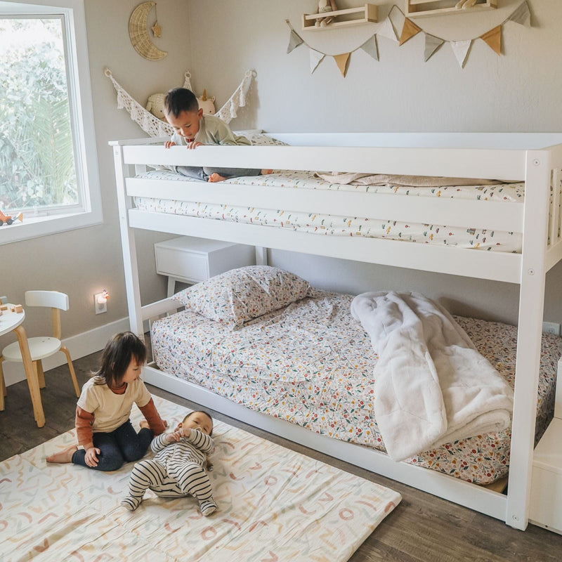 Children's bedroom with white bunk bed and floral bedding, neutral boho nursery decor and bunting, natural light from window and two kids playing on floor mat