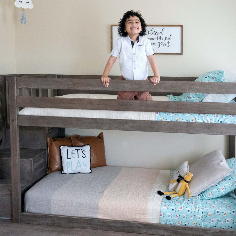 Smiling boy in a gray wooden bunk bed in a kids' bedroom with teal patterned sheets, decorative pillows and nursery wall art
