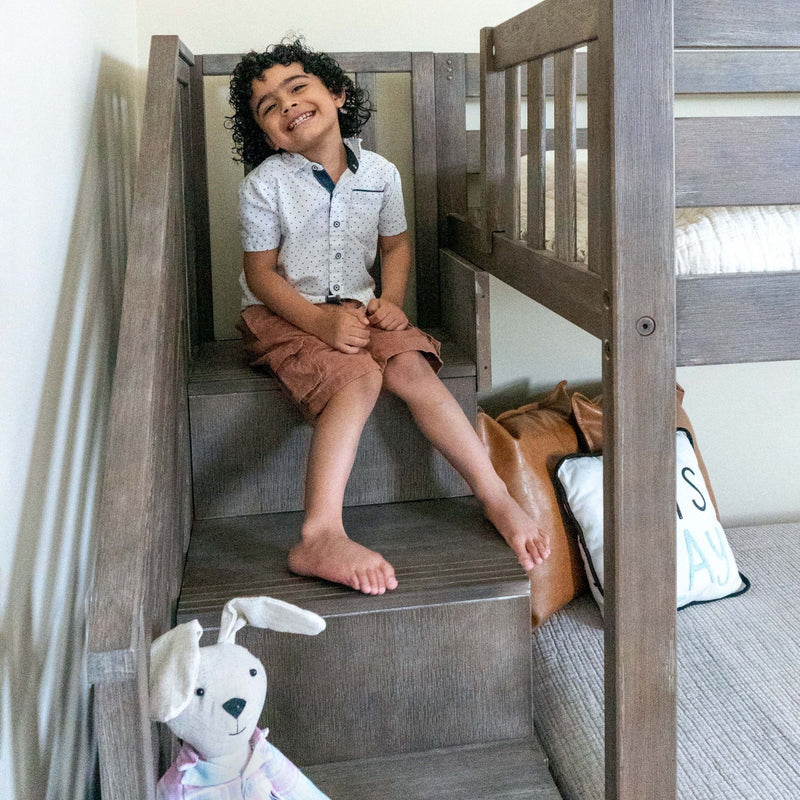 Smiling curly-haired child sitting on wooden bunk bed stairs wearing a white dotted shirt and brown shorts, with a stuffed bunny and decorative pillow in a nursery