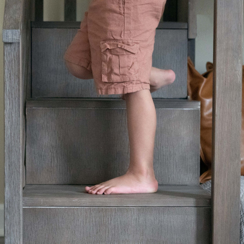 Child's bare feet and legs in salmon cargo shorts climbing gray wooden stairs in a home