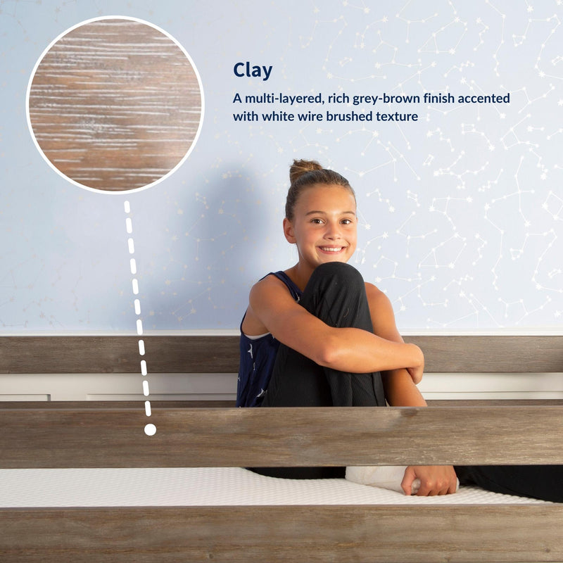 Smiling girl sitting on wooden bed with clay grey-brown wire-brushed wood finish sample and light blue constellation wallpaper