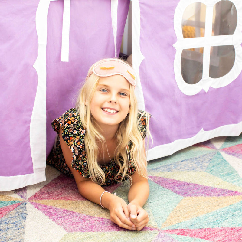 Smiling blonde girl wearing a pink sleep mask peeking from a purple play tent on a colorful rug, kids indoor play and cozy reading nook