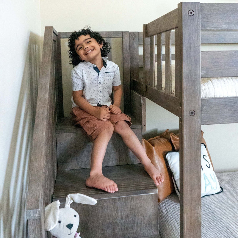 Smiling young boy with curly hair sitting on wooden bunk bed stairs in a bright children's bedroom wearing a white polka-dot shirt and brown shorts, stuffed animal and pillows nearby