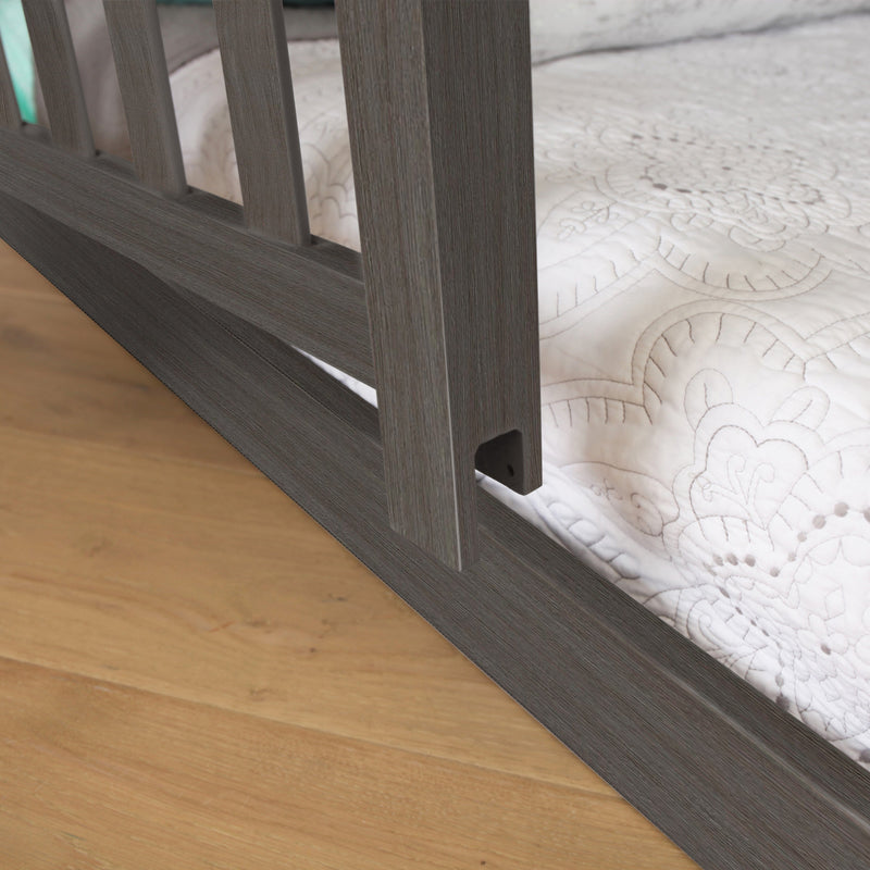 Close-up of gray wood bed frame rail and slats with white quilted mattress cover on hardwood floor