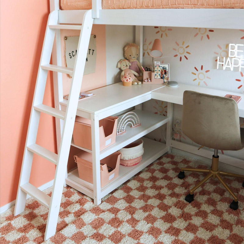 Peach-pink girls' loft bed with white ladder and built-in L-shaped desk, pink storage bins, plush toys, rainbow decor and checkered rug