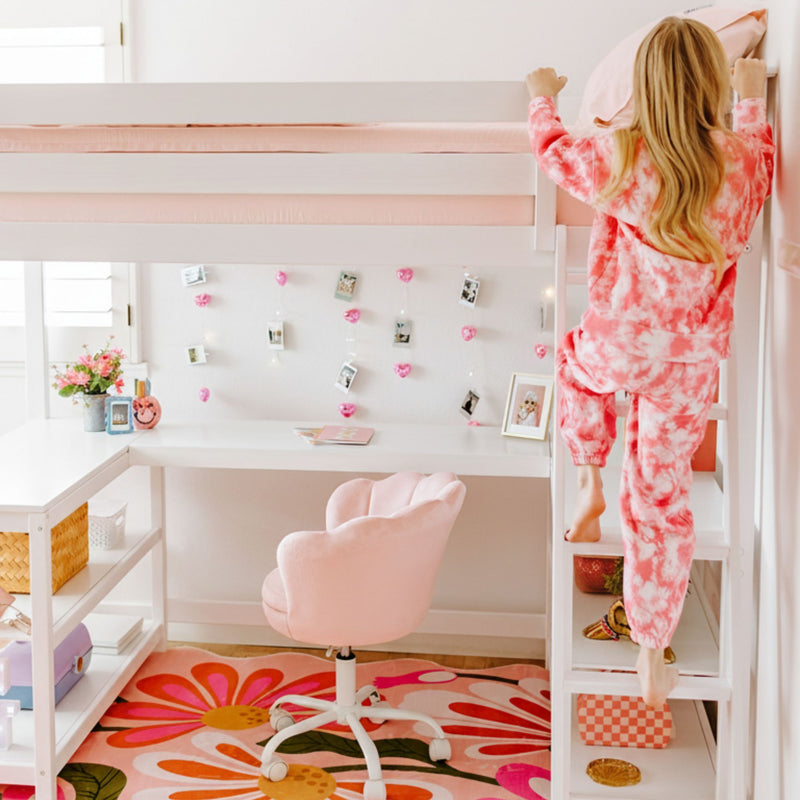 Pink-themed kids loft bed with built-in desk and ladder, pink swivel chair, floral rug and string photo lights, child climbing top bunk