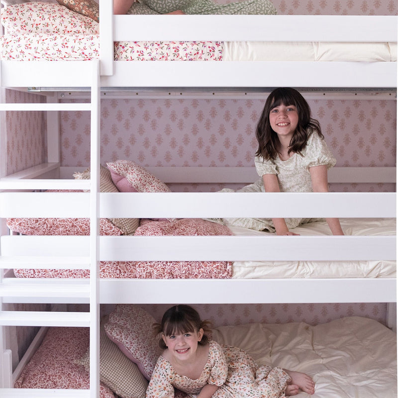 Two smiling girls on white bunk beds with pink floral bedding and patterned wallpaper in a cozy kids' bedroom