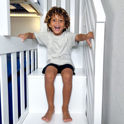 Smiling young boy with curly hair sitting on white bunk bed stairs in a bright modern kids' bedroom