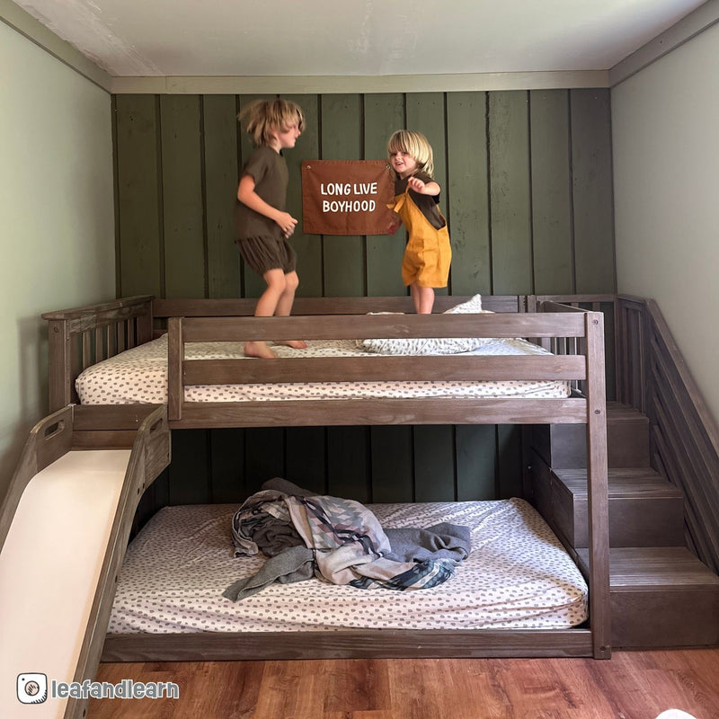 Two children jumping on a wooden loft bunk bed with slide and stairs, green paneled wall and a "Long Live Boyhood" banner, cozy boys' bedroom