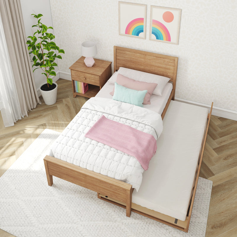 Kids bedroom with wooden single bed and pull-out trundle, pastel pink and mint bedding, wooden nightstand with lamp, potted plant, and rainbow wall art.