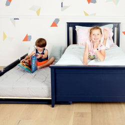 Smiling girl on a navy blue wooden twin bed with pastel bedding and a boy reading on a pull-out trundle mattress in a children's room with geometric wall decals