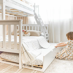 Bright neutral kids bedroom with white wooden bunk bed and pull-out trundle, ladder, cozy quilted bedding, plaid throw and plush toys, child playing on floor — space-saving children's bedroom furniture and nursery decor