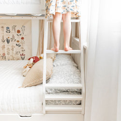 Child's bare feet on a white bunk bed ladder in a bright, neutral nursery with patterned bedding, gingham pillow and botanical wall art