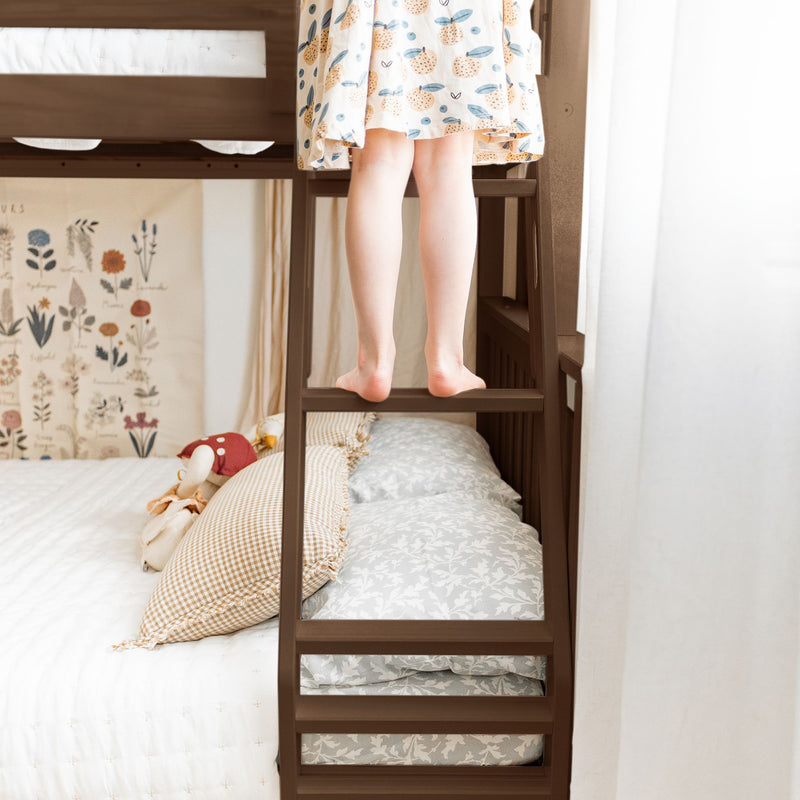 Child on wooden bunk bed ladder in cozy kids bedroom wearing fruit-print dress, floral wall art and layered pillows