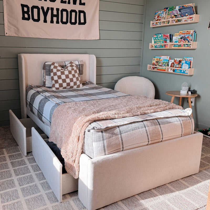Cozy kids' bedroom with beige upholstered twin storage bed and open under-bed drawers, plaid bedding and checkered pillow, soft throw, green shiplap wall with "BOYHOOD" banner and wall-mounted children's book display.