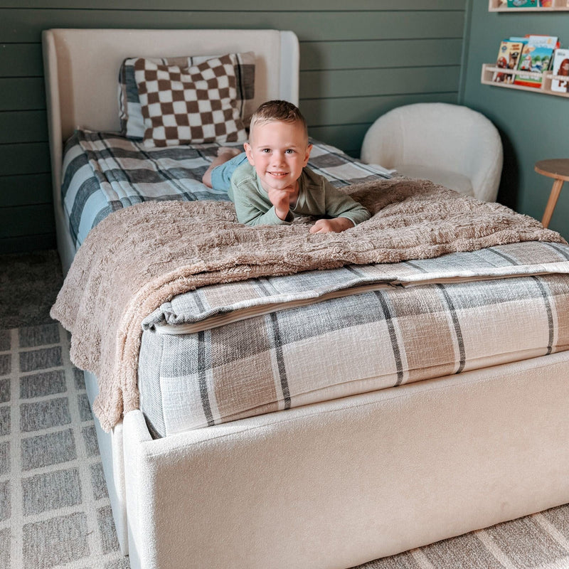 Young child smiling on a cozy bed with plaid bedding in a rustic bedroom setting.