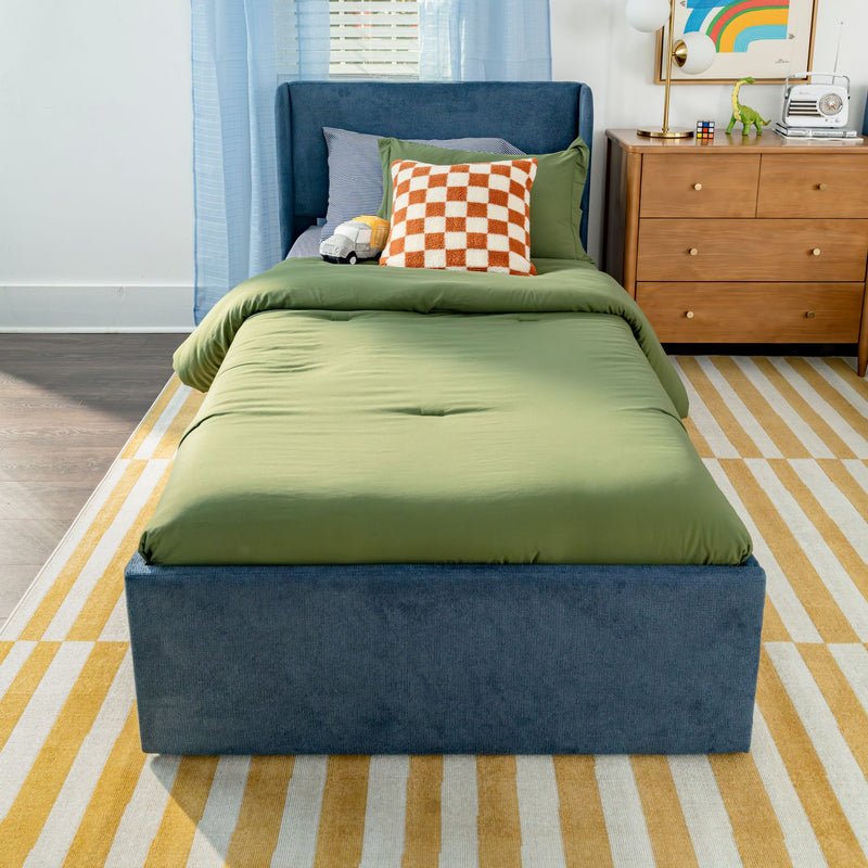 Modern kids bedroom with single blue upholstered bed, green bedding, orange checkered pillow, wooden dresser, striped yellow rug, lamp and toys
