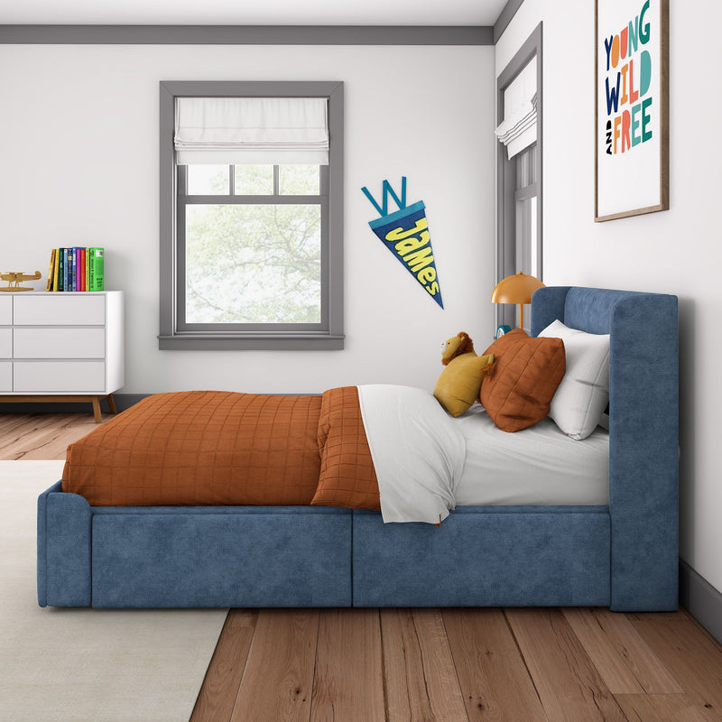 Modern children's bedroom with blue upholstered bed and orange quilt, lion plush toy, white dresser with colorful books, window with Roman shade, personalized pennant and "Young Wild and Free" wall art
