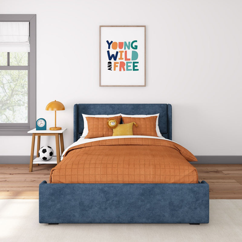 Children's bedroom with blue upholstered bed, orange quilted bedding, framed "Young Wild and Free" wall art, white nightstand with orange lamp and blue alarm clock, and soccer ball on wooden floor