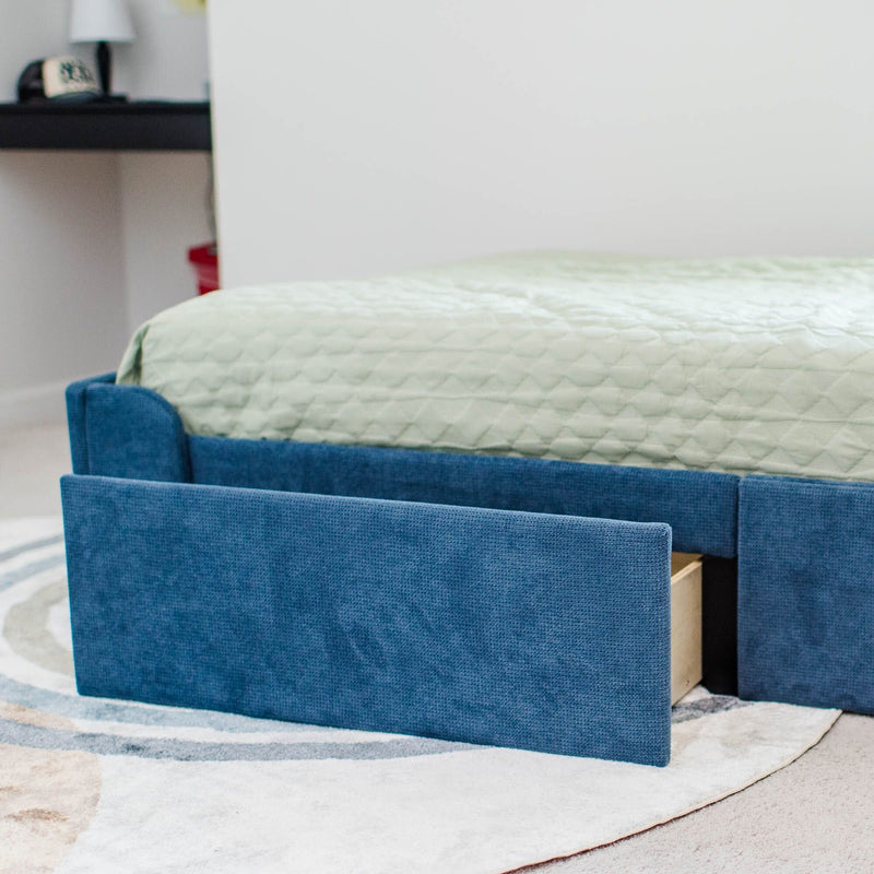 Blue upholstered twin bed frame with open under-bed storage drawer and mint green quilted mattress cover on a patterned rug in a modern bedroom