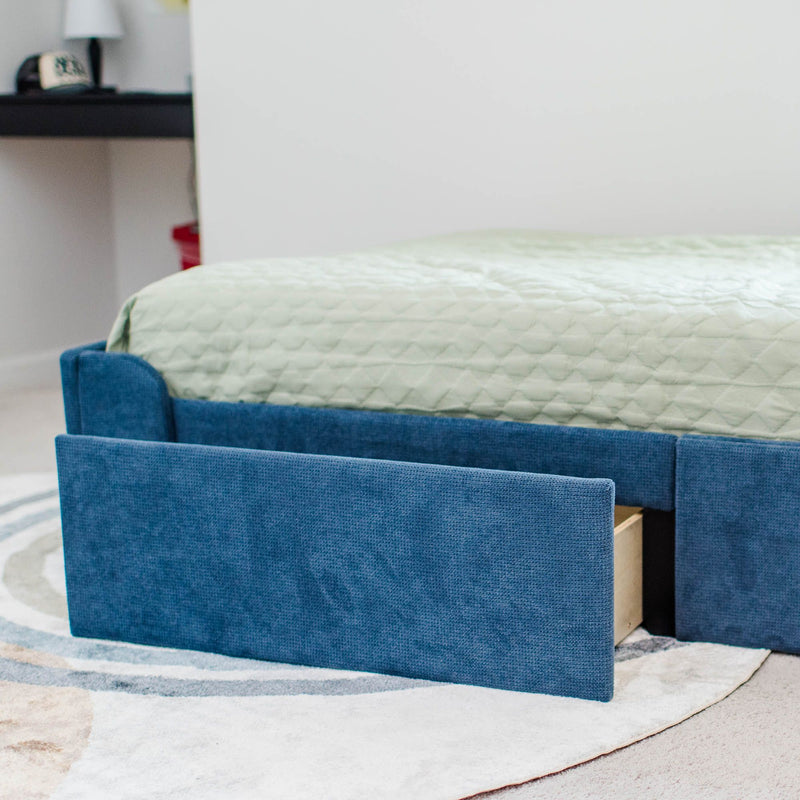 Blue upholstered bed frame with pull-out underbed storage drawer and mint-green quilted bedcover on a neutral circular rug in a modern bedroom