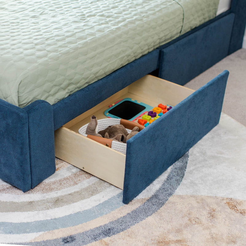 Blue upholstered children's bed with green quilted mattress and open under-bed wooden storage drawers filled with toys on a patterned rug