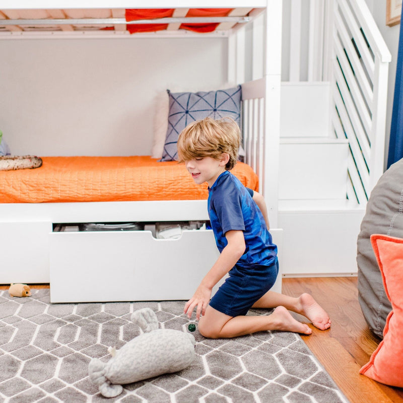 Young boy playing with toy on a gray geometric rug in a bright kids' bedroom featuring a white bunk bed, orange bedding, storage drawer and decorative pillows