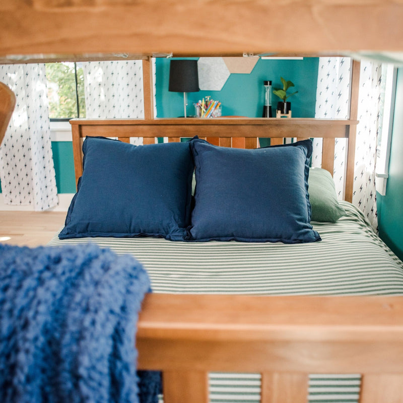 Close-up of wooden bed with two navy pillows on green-and-white striped bedding in a teal bedroom with a black lamp, potted plant, and patterned white curtains