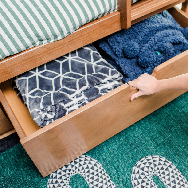 Under-bed wooden storage drawer pulled open revealing folded fleece blankets, patterned throw and green-striped mattress